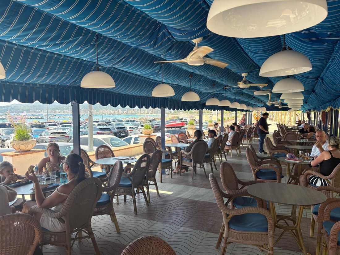 Terraza cubierta de Bell Port con toldo azul, sillas de mimbre y vistas a la bahía de Palamós